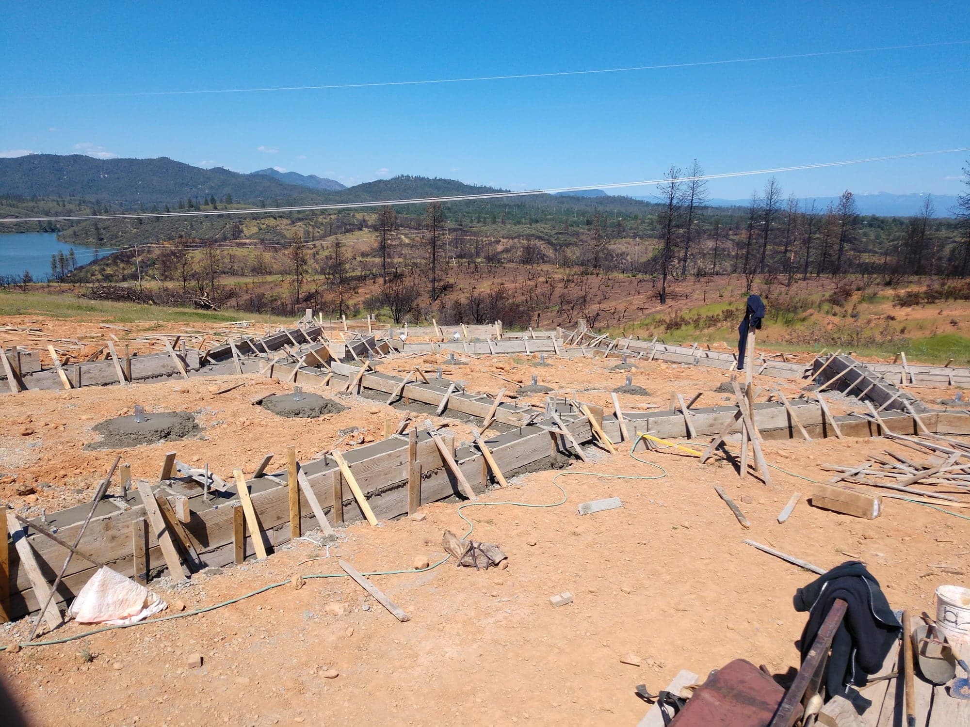 Footing and stem wall forms set out across a ridge above a lake, with burned timber on the far slopes