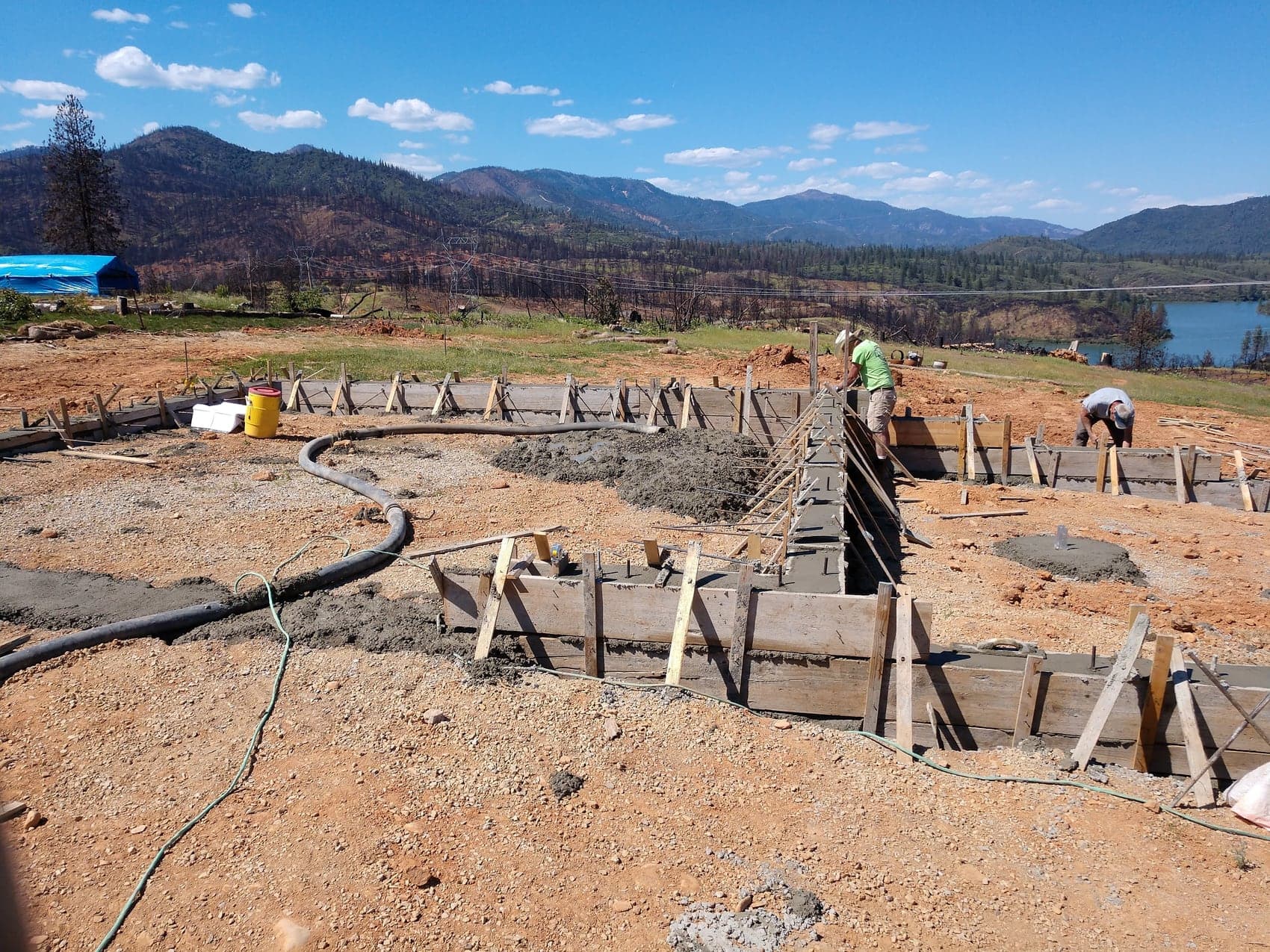 Crew placing concrete into the stem wall forms from a pump line, mountains and lake behind